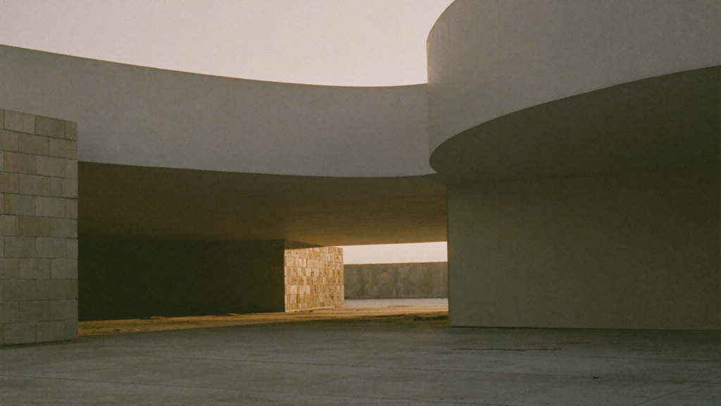 A sweeping curved concrete canopy sheltering a quiet courtyard, with warm golden light falling through the opening beyond a solid stone pillar.