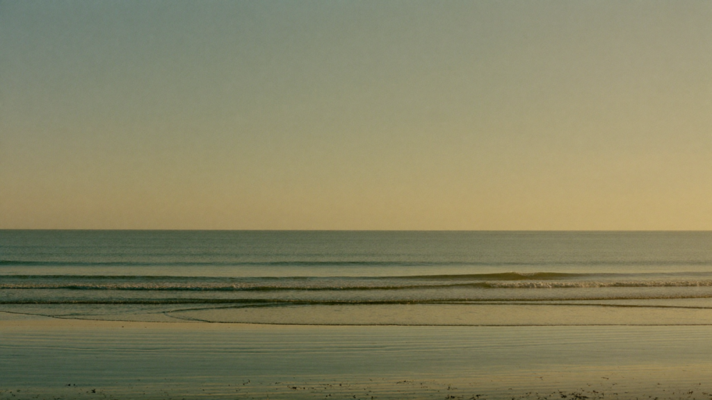 A calm sea meeting a soft golden horizon at first light, seen across still, reflective wet sand under an expansive teal-to-gold sky.