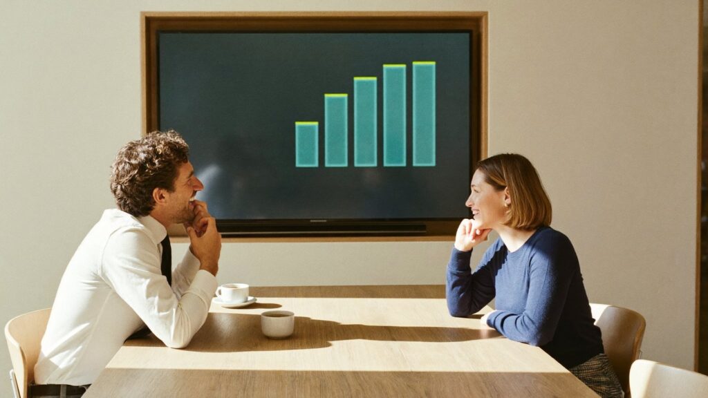 Two people talk across a meeting table beneath a chart screen, asking whether investments rebase on a return to the UK.